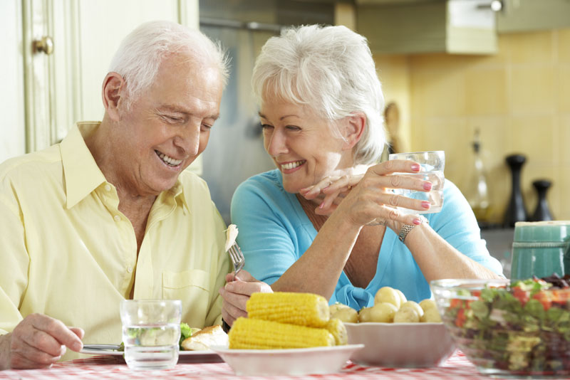 an elderly couple smiling at each other in a well lit dining room while at the dinner table because they have been treated with implant supported dentures.