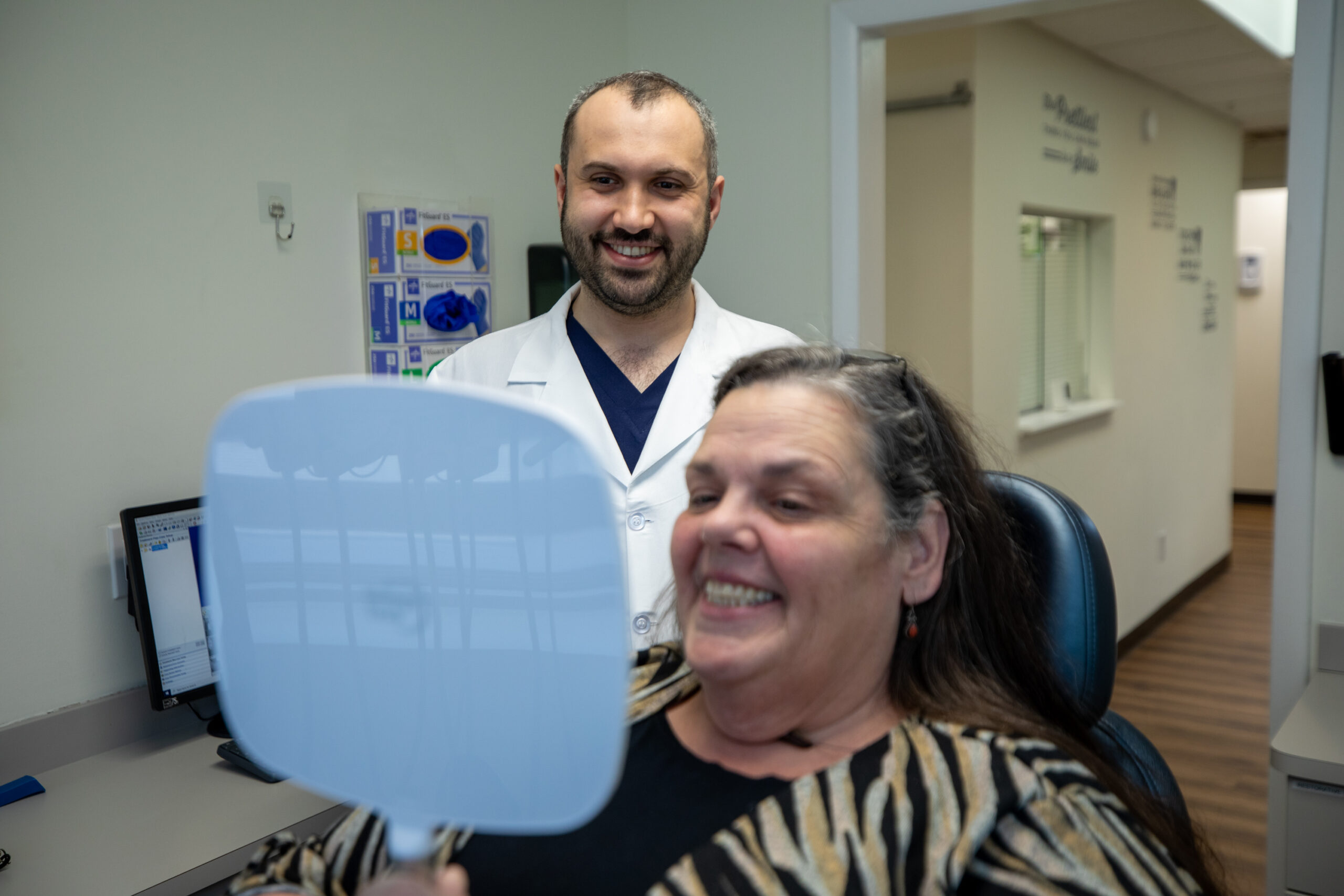 Dr. Basar and his patient smiling at her reflection in a handheld mirror after her zirconia fixed bridge procedure.