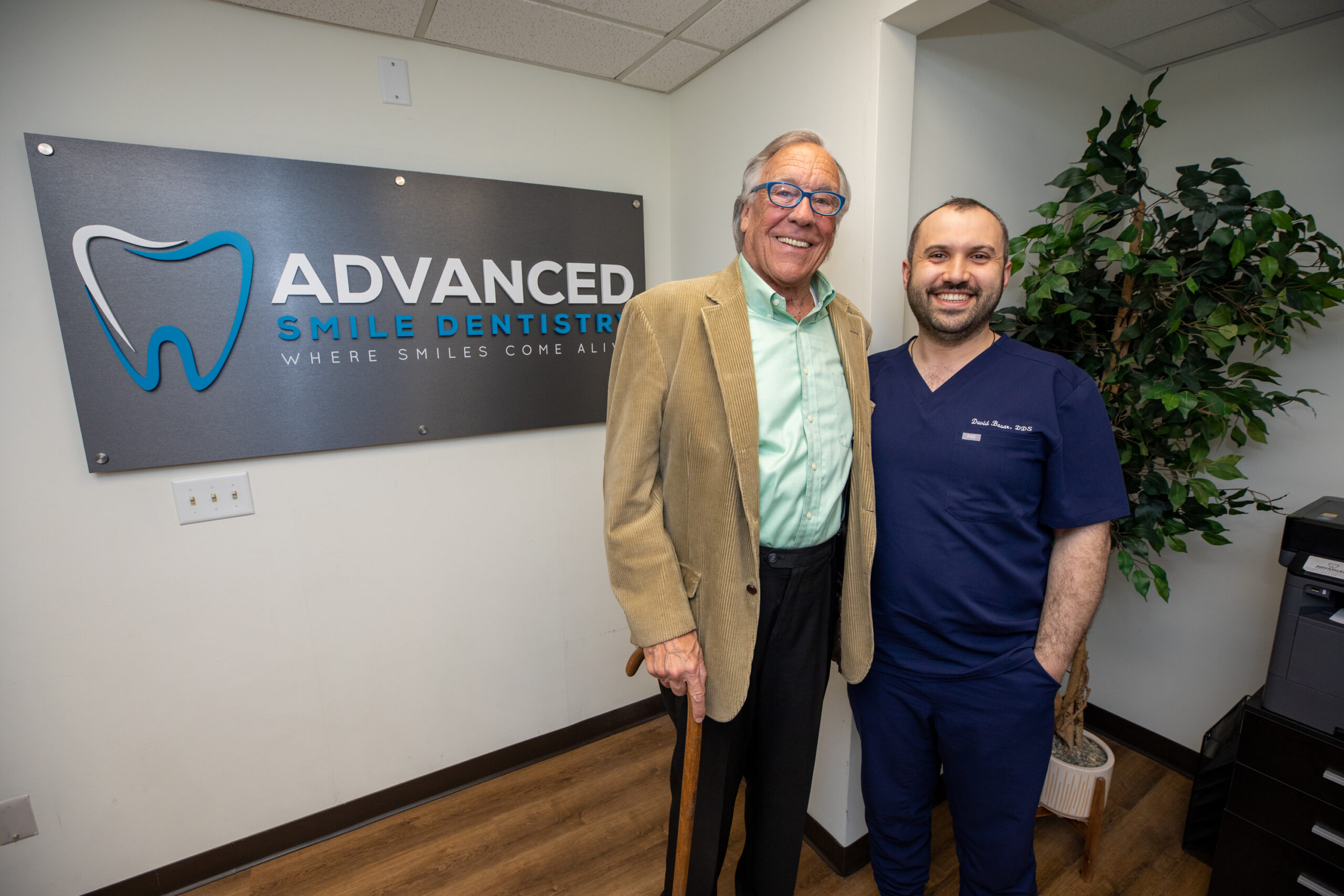 a dental patient and Dr. Basar smiling in front of the practice logo after he has successfully healed from his dental implant surgery.